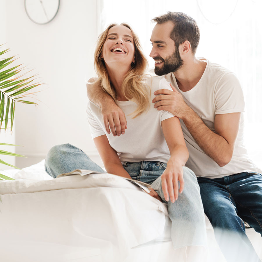 Glückliches Paar sitzt auf einem weissen Bett lacht und freut sich über eine gute Nachricht