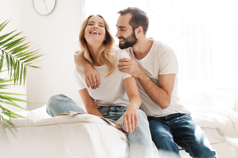 Glückliches Paar sitzt auf einem weissen Bett lacht und freut sich über eine gute Nachricht